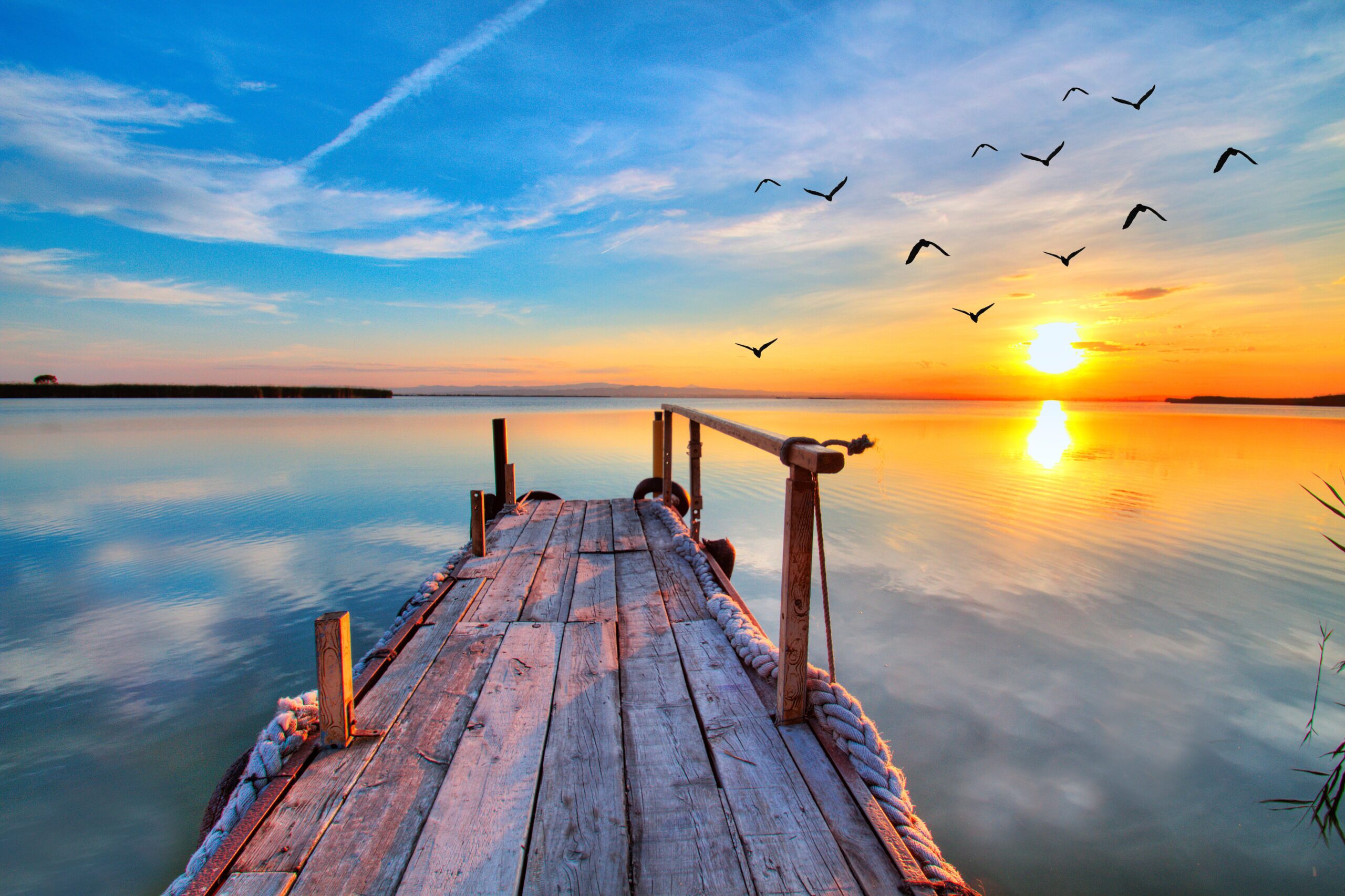 Wooden pier overlooking a serene lake at sunset with birds flying in the sky.