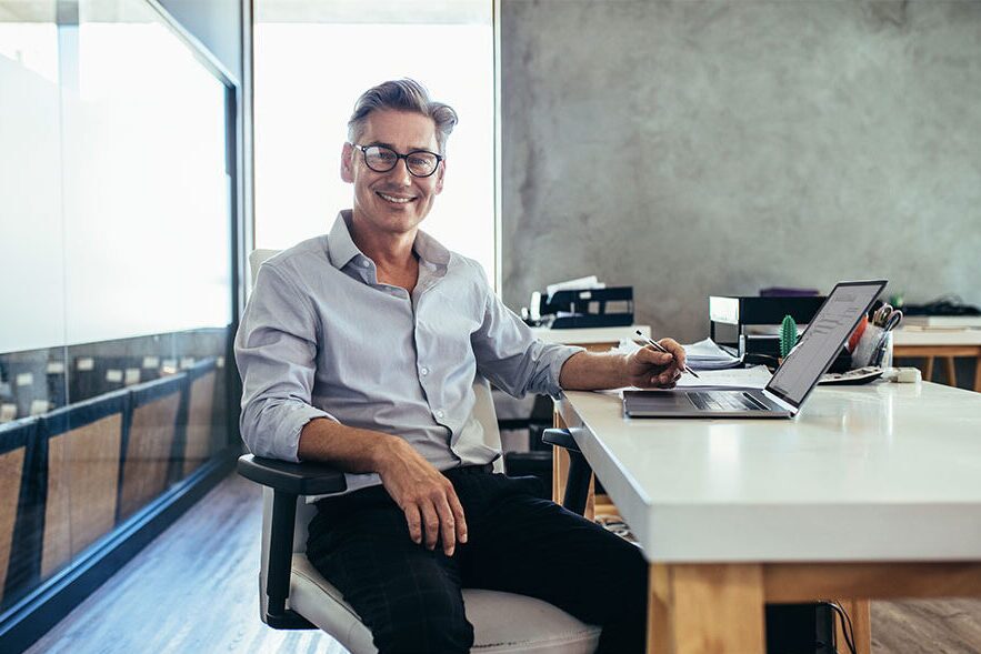 Smiling man in glasses seated at a desk with a laptop in a modern office.