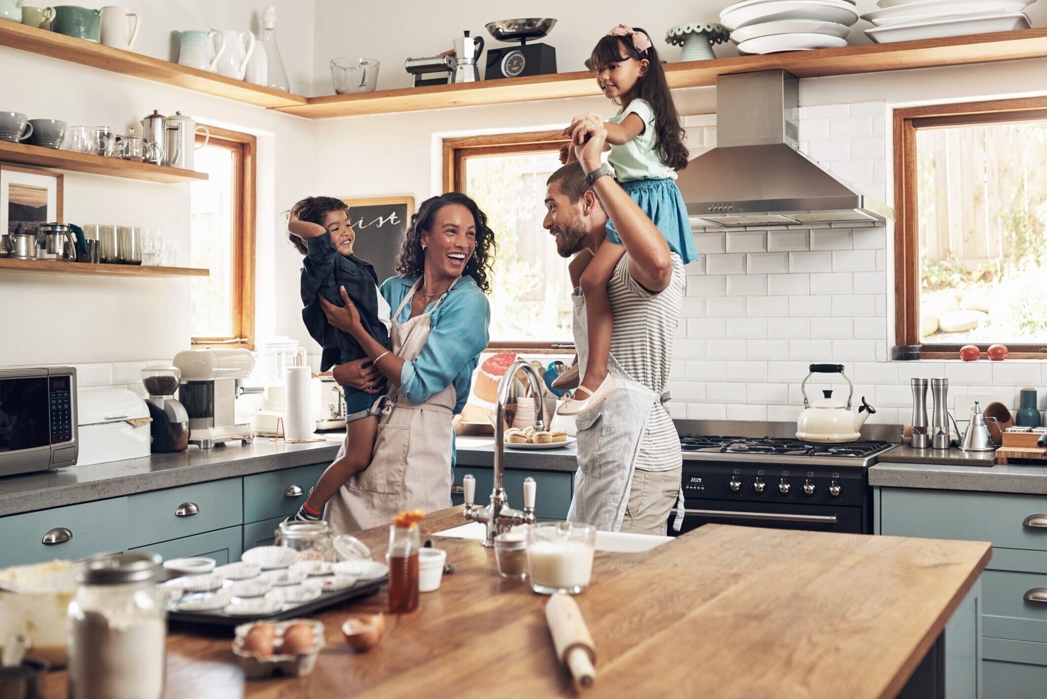 Happy family in kitchen baking with mom holding son and daughter on dad’s shoulders.