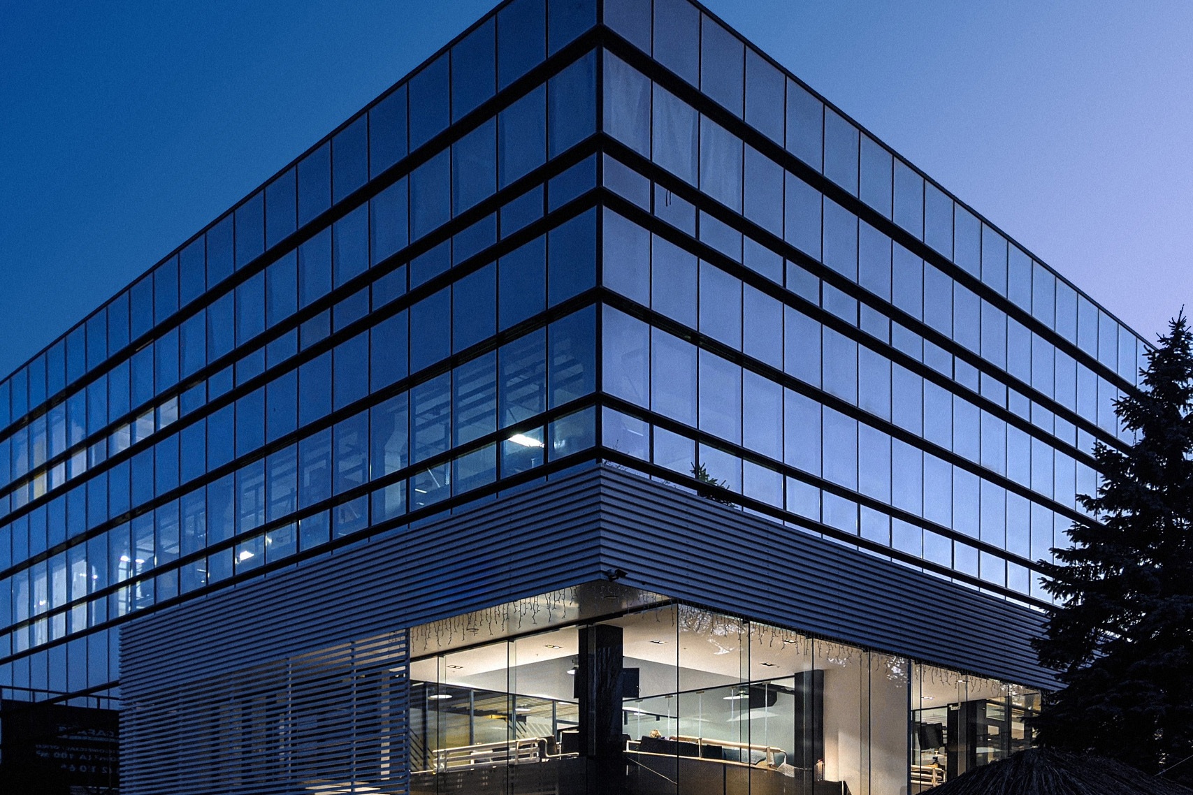 Corner view of a modern glass office building at night, brightly lit with a blue cast.