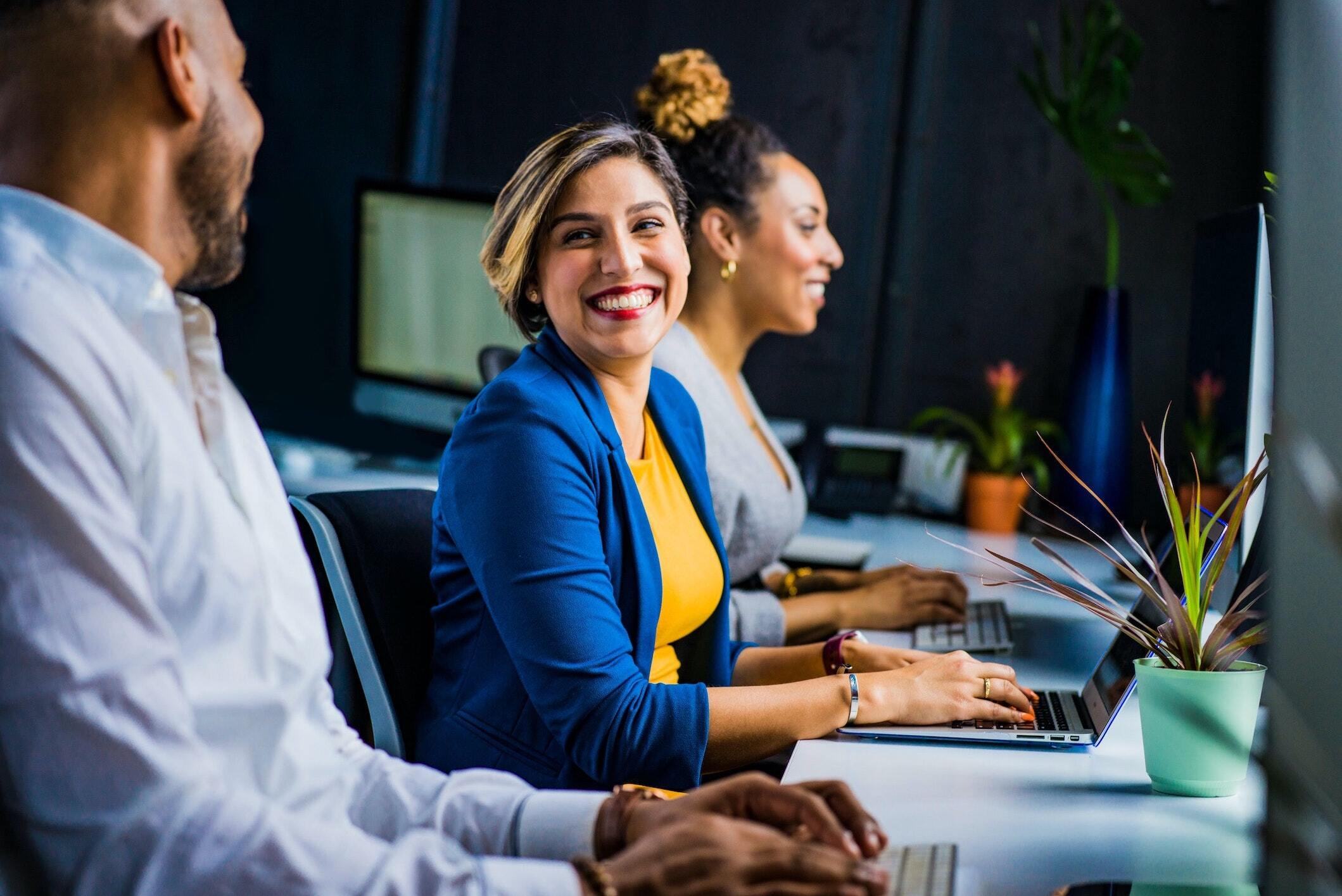 3 smiling employees on computers in a bright workspace.