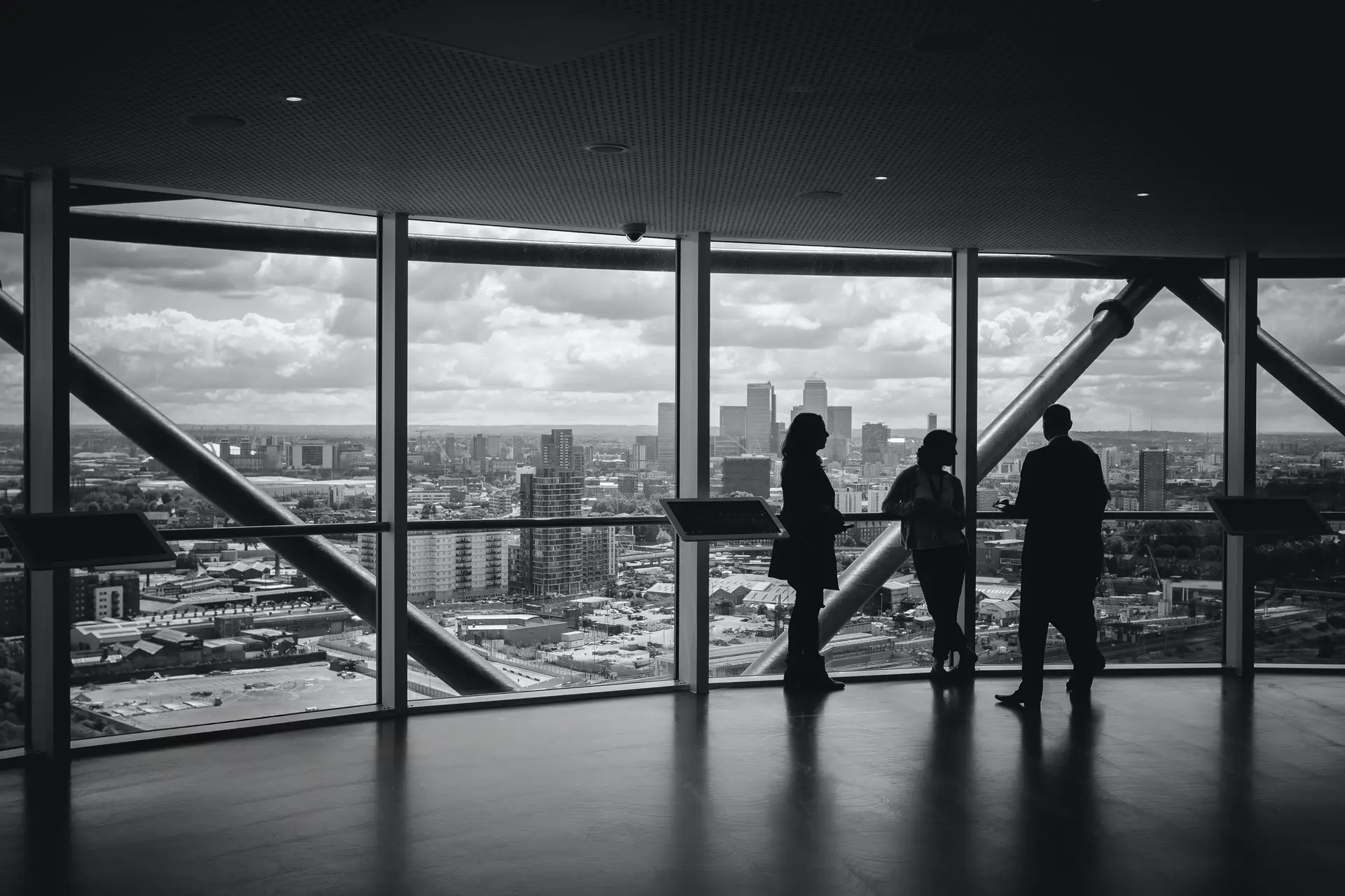 3 silhouetted professionals stand by large windows in a high rise with a cityscape view.